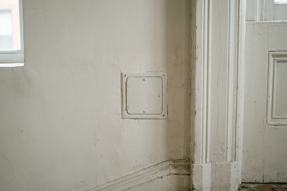 A closed access cover in aged plaster beside preserved doorway trim in an NYC brownstone.