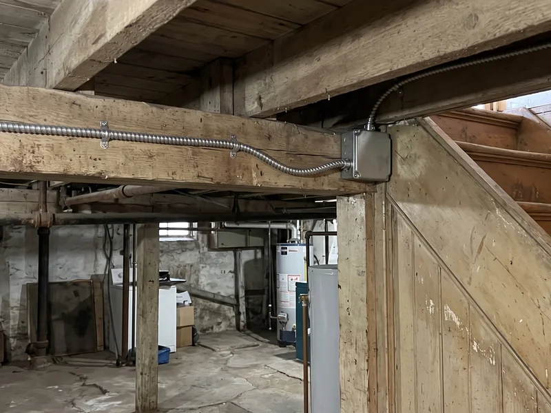 A supported cable along the basement ceiling near an enclosed stair in an older NYC house.